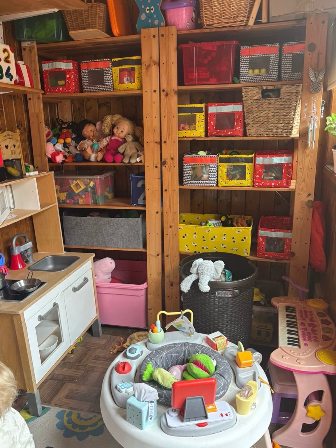 Mary's playroom shed: wooden shelves stacked with toy storage boxes, dolls, a play kitchen and a baby activity centre